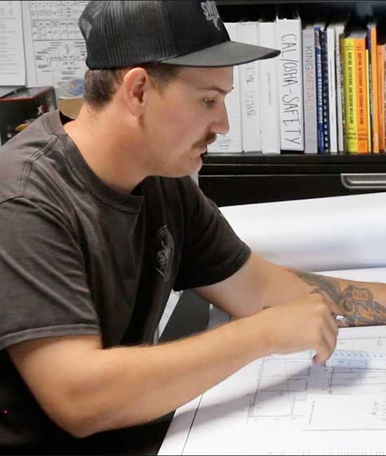 A photo of a mustachioed welder in a ballcap. He's not welding. Imagine that. He's sitting at a drafting table.