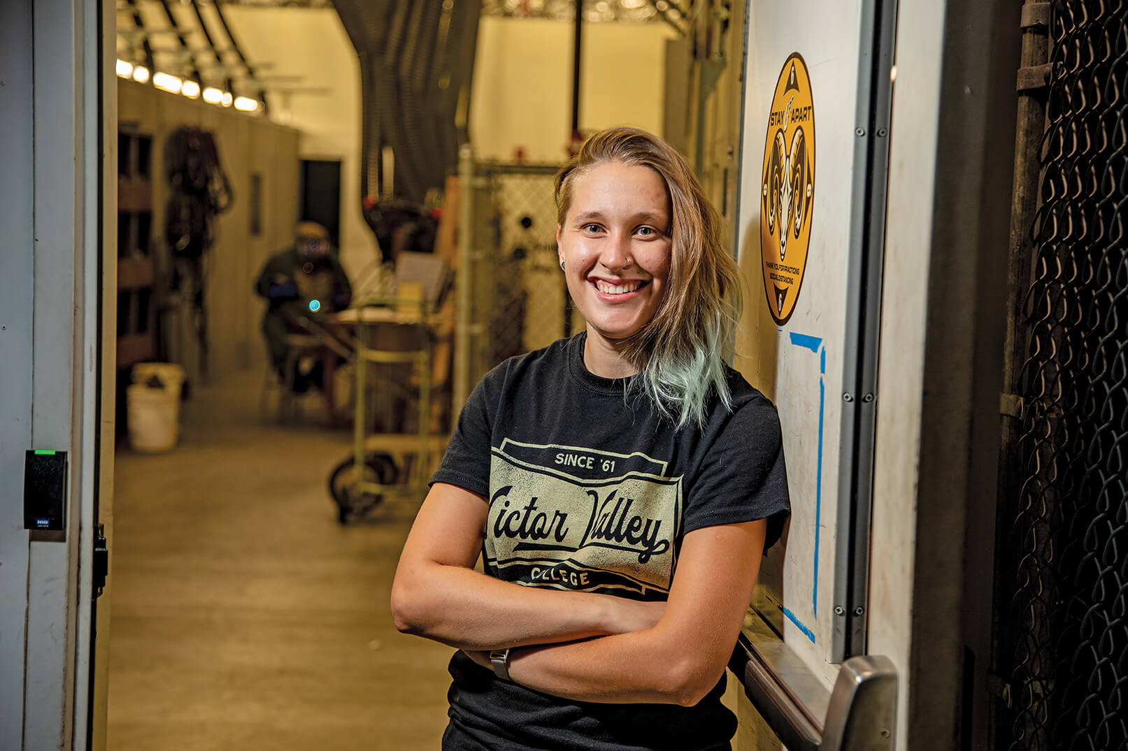 Woman standing in industrial environment, smiling, with arms crossed. She's wearing a Victor Valley College t-shirt.